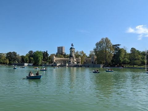 Diversas pessoas navegam em pequenos barcos em um lago de águas esverdeadas. O céu está azul. Atrás do lago, temos uma linha de árvores e um monumento cheio de colunas claras e uma estátua equestre ao centro.