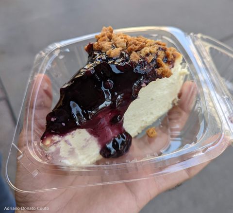 Em um fundo cinza, uma mão segura um pote de plástico com uma fatia de um cheesecake com cobertura roxa.