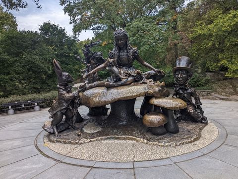 Em uma área aberta rodeada por árvores, temos uma estátua de bronce com alguns cogumelos. Há a escultura de uma menina sobre um dos cogumelos, ladeada por um coelho e um homem com cartola, ambos de bronze.