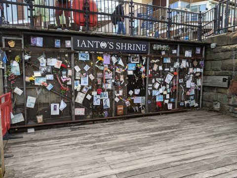 Ao final de uma área forrada em madeira, uma parede escura com muitas mensagens e figuras coladas. Lê-se 'Ianto's Shrine' no topo da parede.".