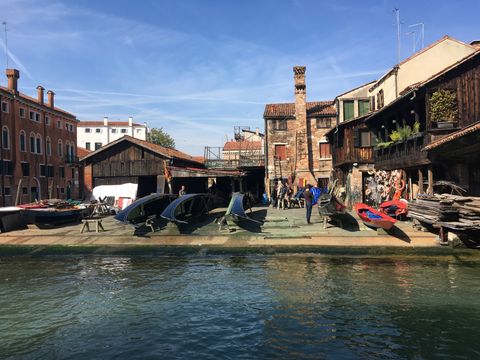 Do outro lado de um canal de águas esverdeadas, vê-se alguns barcos azuis e um vermelho apoiados de lado, em uma área retangular cercada de prédios antigos. É uma oficina tradicional para as embarcações de madeira típicas de Veneza.