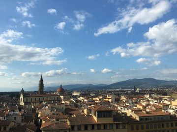 Vista da cidade de Firenze, sob um céu azul cheio de nuvens. A maioria dos prédios tem paredes amarelas. Entre as construções, se destacam a cúpula de uma igreja e uma torre. Ao fundo, montanhas escuras.