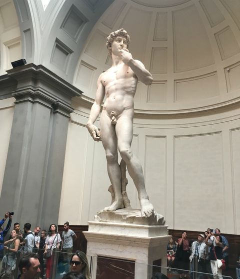 Estátua David di Michelangelo, em destaque em uma área predominantemente branca e cinza. A figura representada, um homem jovem e forte, segura um recipiente raso com a mão esquerda próxima à boca, a mão direita cutucando a coxa direita e perna esquerda apoiada de forma parcialmente flexionada. Em tom sério, é como se observasse algo ao longe, com certo tom de preocupação. Estátua toda branca.