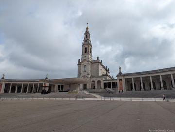 Sob um nublado e cinzento, vemos um espaço cimentado, com colunatas laterais e uma igreja central, com uma torre bem alta.