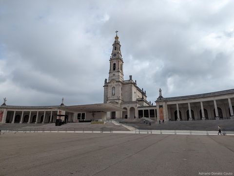 Sob um nublado e cinzento, vemos um espaço cimentado, com colunatas laterais e uma igreja central, com uma torre bem alta.