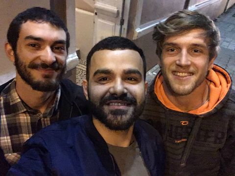 Três rapazes, dois morenos e um loiro, sorriem para uma foto. Ao fundo, um prédio marrom.