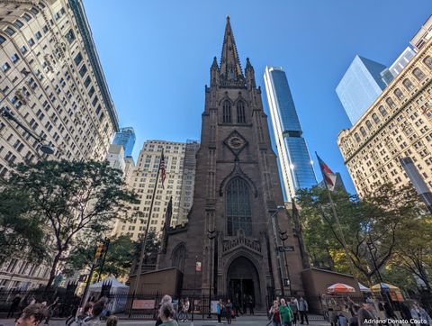 Em uma rua cheia de pessoas e ladeada por muitos prédios altos, vemos uma igreja marrom alta e estreita. Ela tem uma grande porta arqueada no centro e uma única torre.