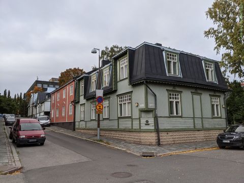 "Vista a partir de uma esquina, temos várias casas de madeira em sequência. A primeira é verde e a segunda, vermelha. As casas possuem dois andares, com janelas grandes."