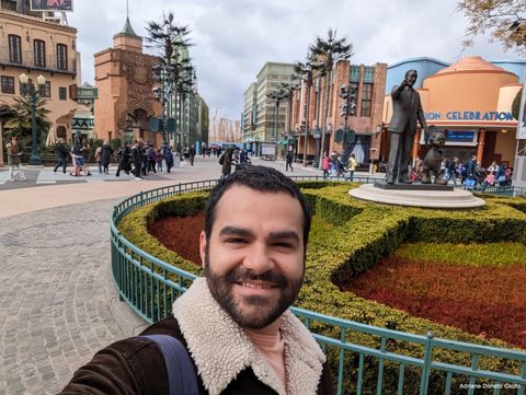 Um rapaz moreno de jaqueta marrom em primeiro plano olha sorrindo para a câmera. Ao fundo, a estátua de bronze de um senhor segurando a mão de Mickey Mouse sobre um trecho de jardim. Ao fundo, prédios alaranjados.