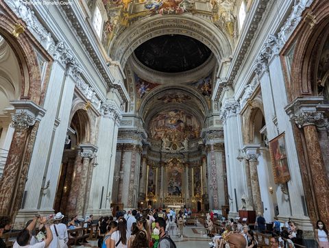Vê-se um grupo de pessoas no corredor central de uma igreja com ricos detalhes esculpidos na paderes, arcos de mármore nas laterais, um domo escuro mais ao fundo e, mais distante, um altar com esculturas e detalhes dourados.