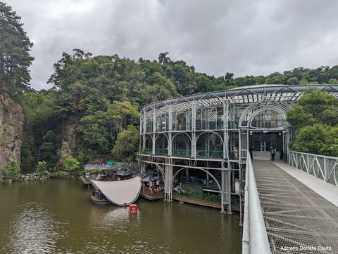 Uma rampa de tubos metálicos claros leva a uma construção do mesmo material. Abaixo, um lago; ao fundo, densa linha de árvores no topo de uma parede de rocha.