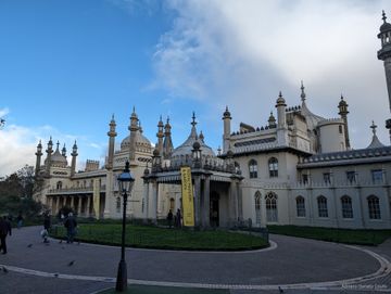 Sob um céu azul e cheio de nuvens, vemos algumas pessoas caminhando em frente a um palácio bege, repleto de cúpulas em um estilo que lembra aquelas do Taj Mahal.
