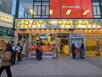 Em frente a uma fachada amarela em que se lê Gray's Papaya, de uma loja de hot dogs, algumas pessoas soboreiam a iguaria e outras caminham pela calçada.