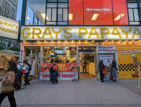Em frente a uma fachada amarela em que se lê Gray's Papaya, de uma loja de hot dogs, algumas pessoas soboreiam a iguaria e outras caminham pela calçada.