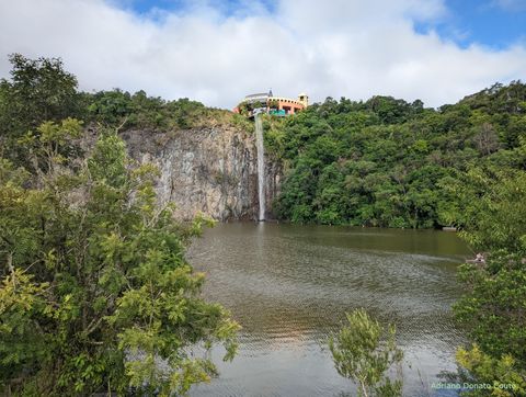 Em primeiro plano, um grande lago. Ao fundo, um paredão de rocha e uma cortina d'água No topo, uma construção amarela.