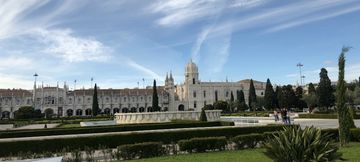 Extensa área gramada, com algumas árvores e um espelho d'água. Ao fundo, uma longa fachada construída de cor clara, sendo uma parte  de uma igreja e a outra parte de um mosteiro. Por cima, um céu azul com muitas nuvens ralas.