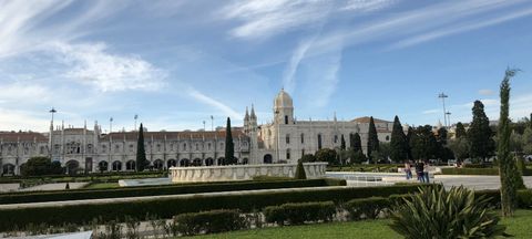 Extensa área gramada, com algumas árvores e um espelho d'água. Ao fundo, uma longa fachada construída de cor clara, sendo uma parte  de uma igreja e a outra parte de um mosteiro. Por cima, um céu azul com muitas nuvens ralas.