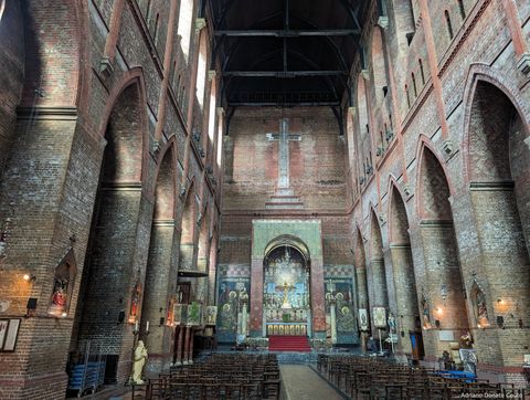 'Vemos o interior de uma igreja muito alta, ladeada por arcos igualmente alongados verticalmente. O revestimento é todo de tijolos avermelhados, ornados com pinturas com detalhes dourados e uma cruz enorme.'