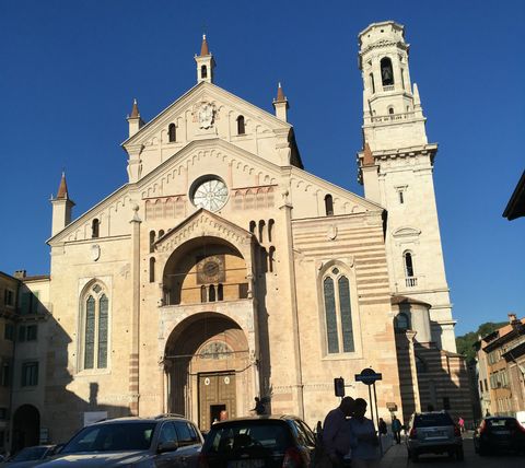 Um céu azul sem nuvens no topo da imagem, com uma catedral em tons claros de marrom logo abaixo. No topo da catedral, ao centro, uma rosácea. Embaixo, uma área de sombra com carros estacionados e algumas pessoas.