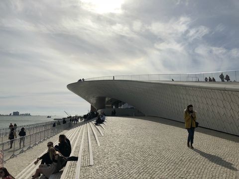 Uma construção coberta por placas claras avança em formato de onda sobre uma escadaria. Ao fundo, temos o rio e um céu coberto por nuvens.