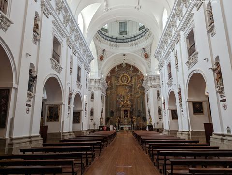 Um longo corredor com piso de madeira avermelhada, ladeado por bancos de madeira, com um altar todo trabalhado e com peças douradas no fundo. Em volta, uma série de arcos em paredes brancas, com o topo todo trabalhado em relevo da mesma cor.