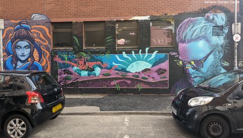 Entre dois carros estacionados, vemos uma parede de tijolos vermelhos quase toda coberta com grafites em tons frios.