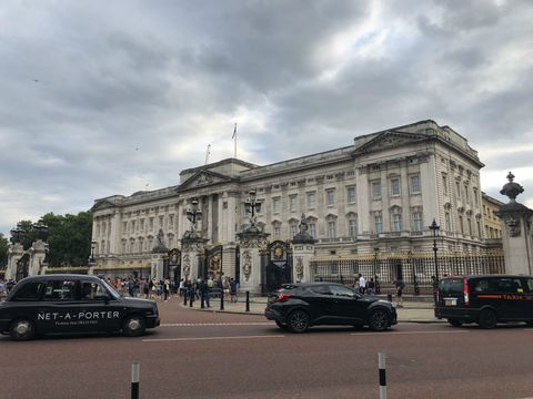 Sob um céu carregado de nuvens e logo após uma via de asfalto avermelhado, uma cerca preta de ferro com detalhes em dourado separa o público de um grande palácio claro, com um número interminável de janelas. Na rua, há três veículos pretos passando.