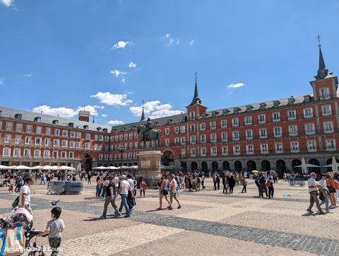 Uma praça repleta de pessoas caminhando em diferentes direções. Ao centro, uma estátua equestre. Ao fundo, rodeando a praça, um grupo de prédios de paredes vermelhas e incontáveis janelas.