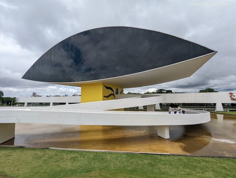 Sobre um espelho d'água, vê-se um conjunto de rampas de concreto pintadas de branco. Ao centro, surge uma pilastra com uma construção em formato de elipse no topo.