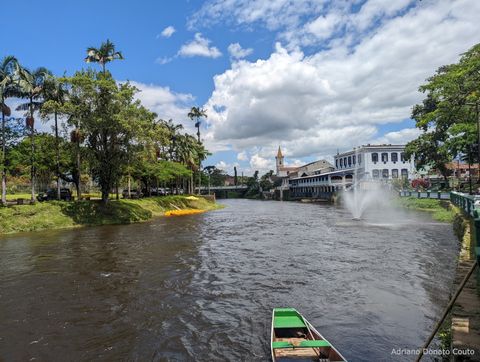 Vê-se um rio que parte do primeiro plano e vai até o fundo. De um lado, palmeiras; do outro, um casarão branco de detalhes azuis e uma igreja.