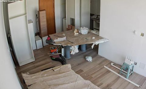 'Uma sala branca de piso amadeirado com móveis cobertos por papelão, com uma série de armários brancos e de madeira espalhados, que estão parcialmente montados.'