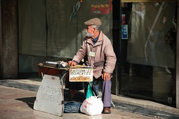 Em frente a um local cujas vitrines estão cobertas com papel, um senhor de cabelos grisalhos está sentado. à sua frente, uma banca de madeira. Há duas placas que anunciam o serviço prestado: plastificam-se documentos.