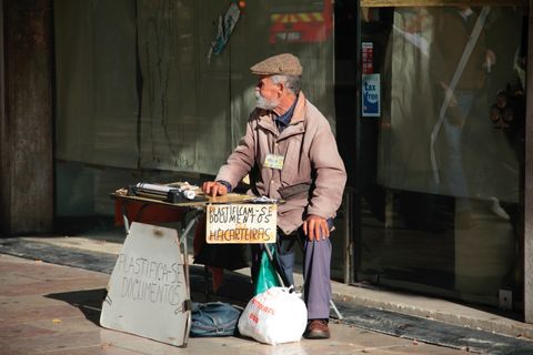 Em frente a um local cujas vitrines estão cobertas com papel, um senhor de cabelos grisalhos está sentado. à sua frente, uma banca de madeira. Há duas placas que anunciam o serviço prestado: plastificam-se documentos.