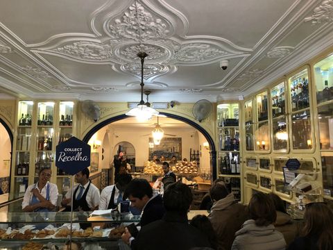 Interior de um estabelecimento antigo, com paredes amarelas cheias de vitrines e o teto branco, com detalhes em relevo e alguns pendentes de luz. Há pessoas atrás do balcão e clientes do outro lado. À esquerda, uma placa diz "Recolha" (coleta, em português de Portugal).