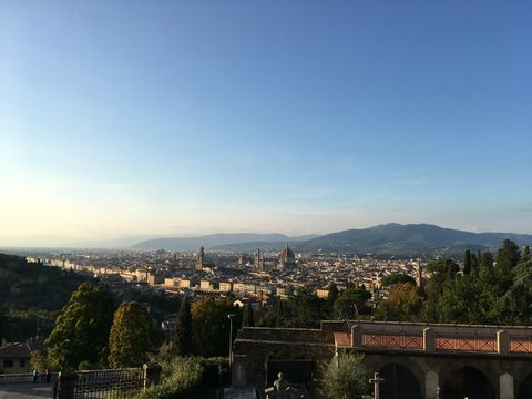 Vista de Florença a partir de um monte. Em primeiro plano, uma passarela e algumas árvores.Logo após, a cidade de Florença, com algumas cúpulas e torres em destaque. De fundo, algumas montanhas, já meio apagadas pela distância.