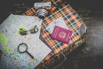 Sobre um piso manchado, há uma maleta xadrez, em tons de laranja, preto e branco. Sobre a maleta, um par de óculos de sol, lupa, mapas, uma câmera fotográfica retrô, cédulas de dinheiro e um passaporte italiano (vermelho).