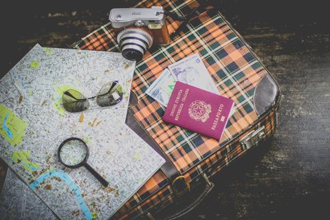 Sobre um piso manchado, há uma maleta xadrez, em tons de laranja, preto e branco. Sobre a maleta, um par de óculos de sol, lupa, mapas, uma câmera fotográfica retrô, cédulas de dinheiro e um passaporte italiano (vermelho).