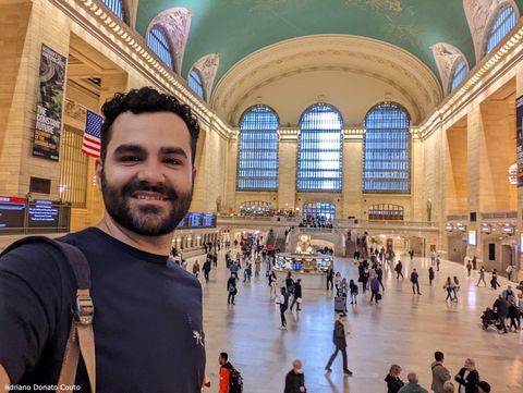 Em primeiro plano, um rapaz de camiseta azul tira uma selfie. Ao fundo, um bom número de pessoas caminha por uma grande sala ricamente decorada. O teto é azul, com desenhos dourados de constelações.