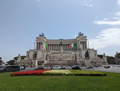 Em primeiro plano, um gramado com a bandeira tricolor italiana feita de flores Ao fundo, uma grandiosa construção bege, com esculturas escuras ornamentando as laterais e o centro. Em um nível superior da construção, a fachada é toda de colunas.