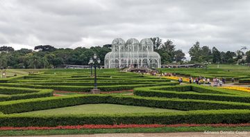 Sob céu azul com muitas nuvens, temos um diagrama de arbustos à frente e um palácio de cristal ao fundo, todo com armação metálica branca. Muitas pessoas passeiam pelo local.