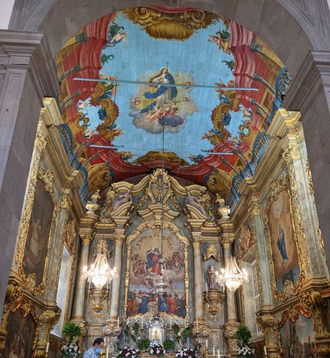 "Destaque para o interior de uma igreja antiga, com foco para a área atrás do altar. No teto, em madeira, uma pintura de Nossa Senhora; nas paredes, um trabalho muito detalhado que mistura molduras douradas e pinturas religiosas."