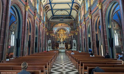 Vê-se uma igreja a partir do fundo, com seu corredor no meio. Ladeado por colunas avermelhadas com o topo dourado, o teto é bege com linhas azuis. Há muitas cores e detalhes. Algumas pessoas estão sentadas nos bancos da igreja.