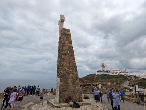 'Há um monumento ao centro da imagem, em formato de torre. Ele tem uma cruz de concreto no topo. Em segundo plano, à esquerda, pessoas observam o mar. Do lado direito, no fundo, temos algumas construções e um farol.'
