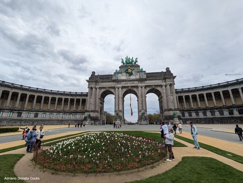 Sob céu nublado e cinza, uma área de jardim em primeiro plano, com algumas pessoas; ao fundo, um arco triunfal com três vãos.