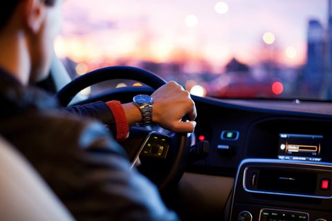 Visão da parte da frente de um veículo a partir do banco de trás. Um homem de jaqueta de couro e relógio com pulseira de metal está com a mão firme ao volante. À direita, destaque para o painel do carro. O vidro da frente traz um exterior desfocado.