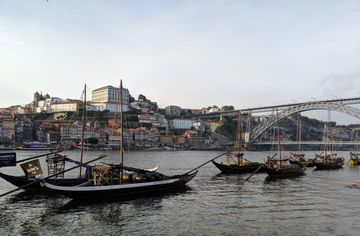 Vista a partir das margens do rio Douro, quase ao nível da água. Algumas embarcações em primeiro plano. Ao fundo, uma colina cheia de construções antigas.