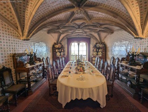 'Sob um teto abobadado todo forrado em azulejos, temos uma enorme mesa de jantar com pratos e talheres, além de um centro de mesa decorado. A mesa é ladeada por cadeiras, aparadores e outros móveis de madeira escura.'