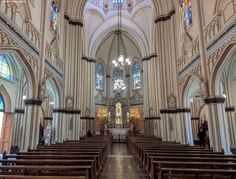 Vê-se uma igreja a partir do fundo, com seu corredor no meio. As paredes possuem muito relevo e alguns arcos, mas a cor predominante é o bege. Há lustres no teto, além de bancos escuros de madeira abaixo.