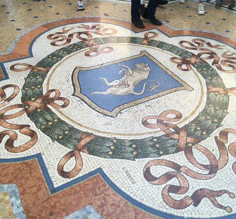 Mosaico no chão de uma galeria, com um brasão azul com um touro em destaque. Em volta, o desenho de uma guirlanda verde com fitas vermelhas.