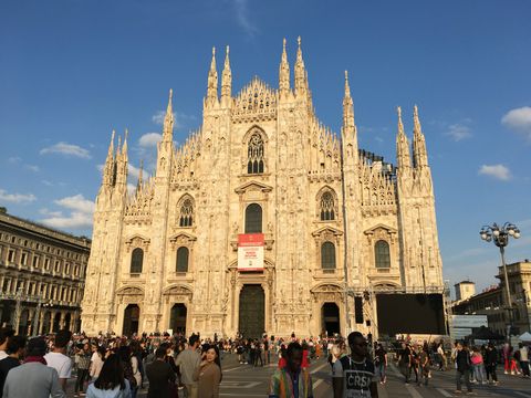 Sob um céu azul com algumas nuvens, um grupo grande de pessoas passeia em frente a uma majestosa catedral.